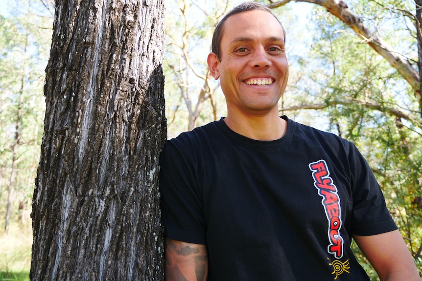 An Indigenous man in a black shirt with the Aboriginal flag stands and looks at the camera.