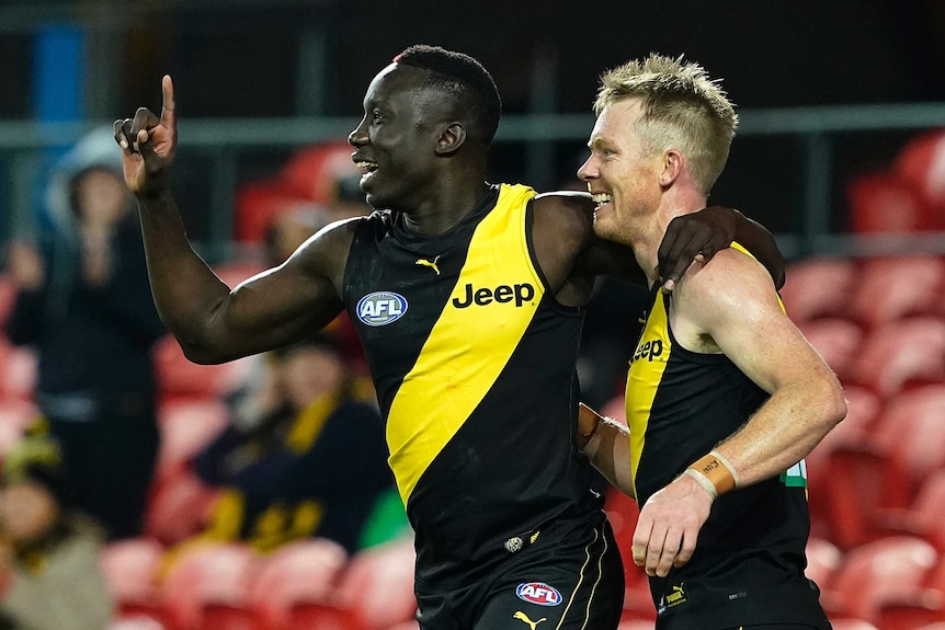 Two AFL teammates celebrate a goal, one raising his finger in salute, the other smiling.