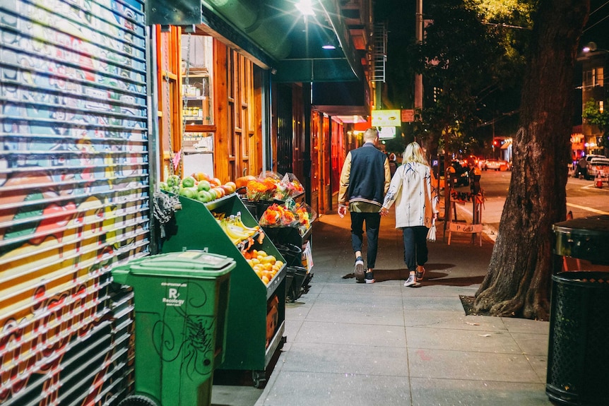 A man and a woman walk down a street at night holding hands.