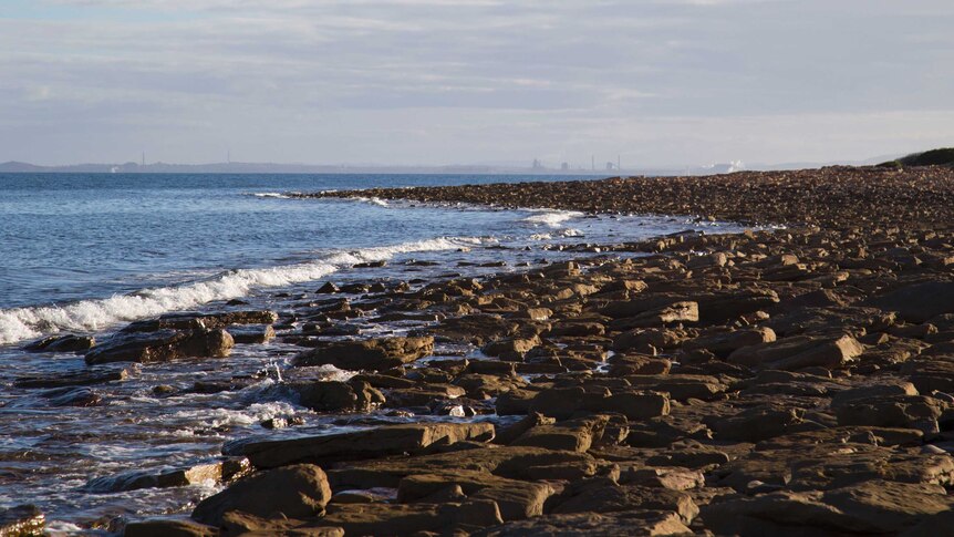 small waves crash on a rocky shore line, factories and industry are in the distance on the horizon