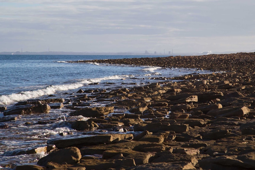 small waves crash on a rocky shore line, factories and industry are in the distance on the horizon
