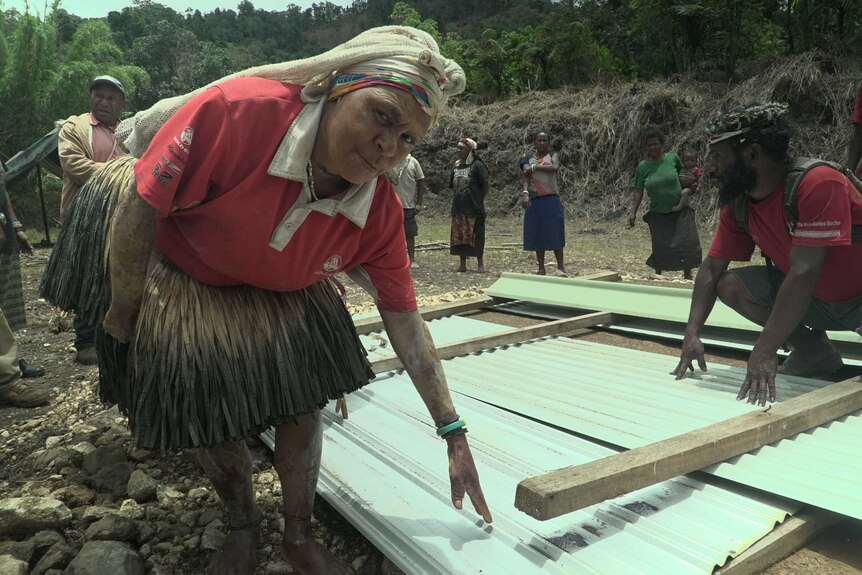 An older woman crouches by corrugated sheeting laid across the ground