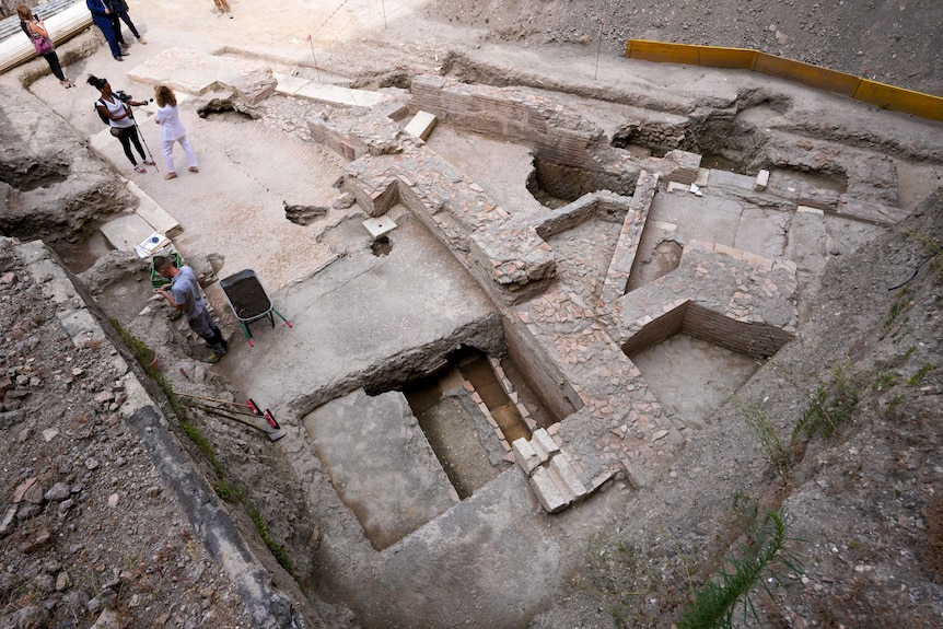 An archaeological dig site with ruins visible and deep holes and people standing around 