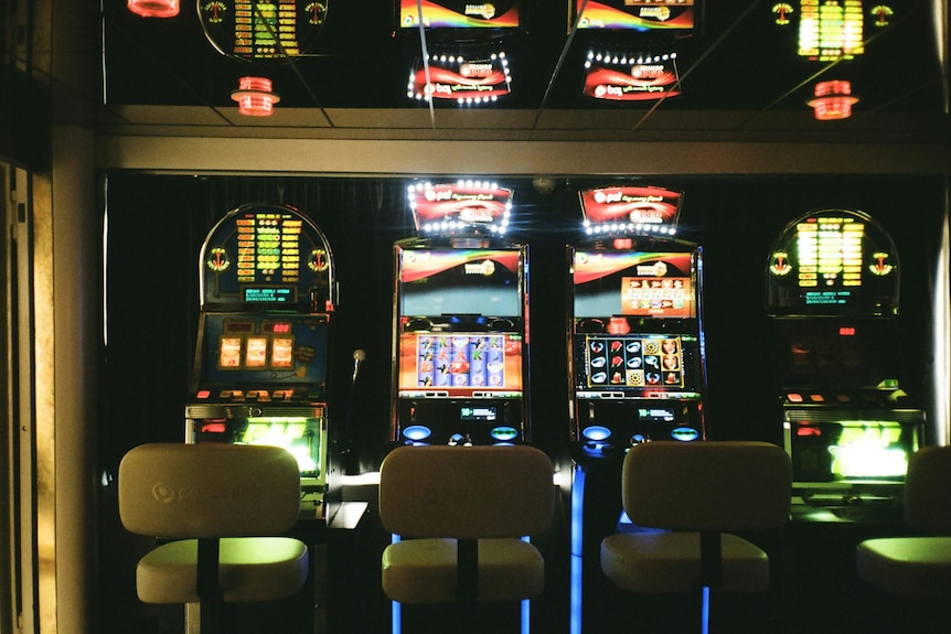 Three white stools in front of three poker machines lit up in a dark gambling room.