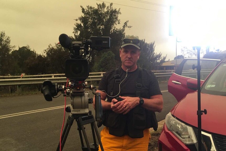 Nelson standing next to camera wearing protective clothing with smoky sky in background.