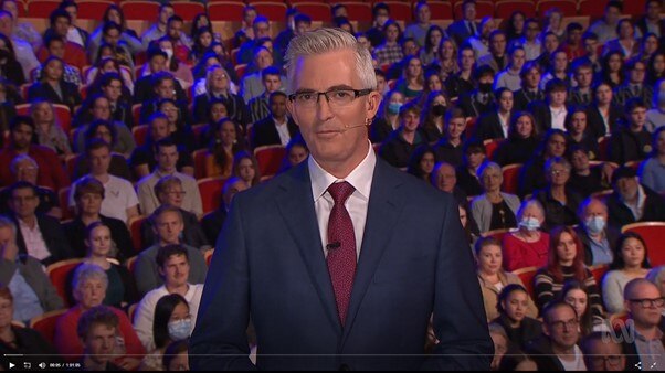 Man in a suit standing in front of a large audience in a TV studio.