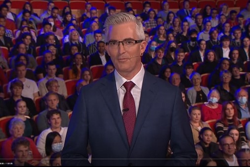 Man in a suit standing in front of a large audience in a TV studio.