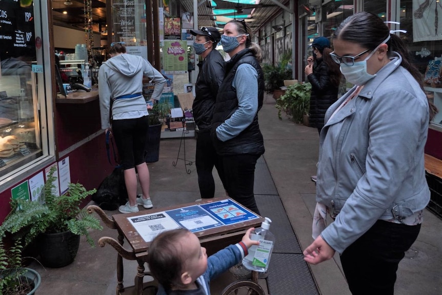 Several people are standing outside a cafe, wearing masks.  A small child holds up a disinfectant bottle