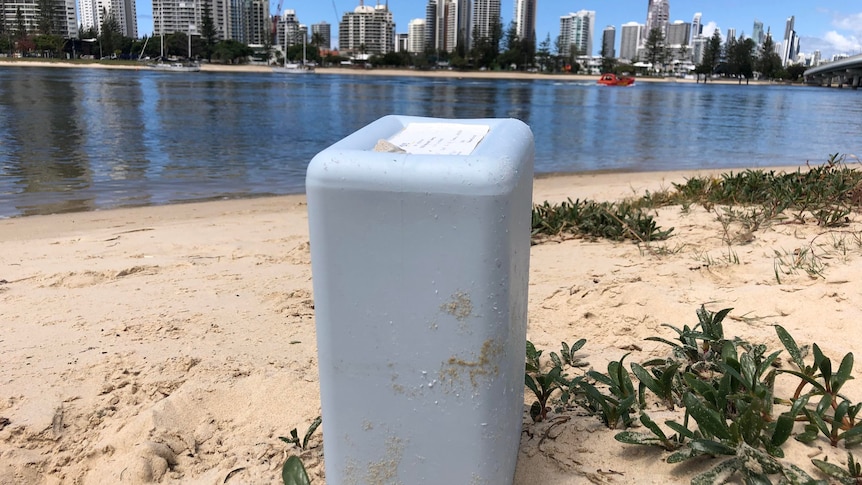 A light blue container sits on sand with the beach behind