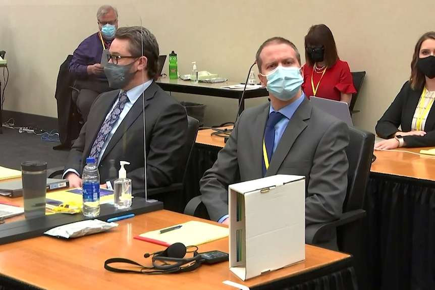 A middle-aged man in grey suit and face mask sits beside another man in dark suit in courtroom.