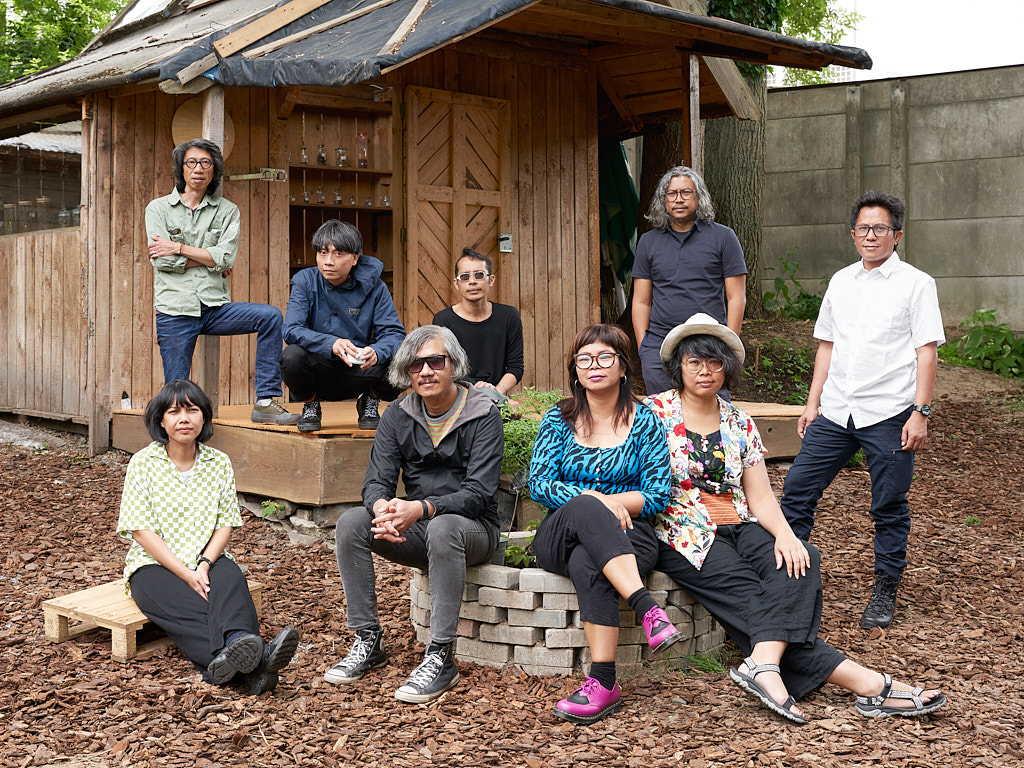 A group of nine Indonesian artists variously sit and stand on wooden benches and stones outside a wooden shack.
