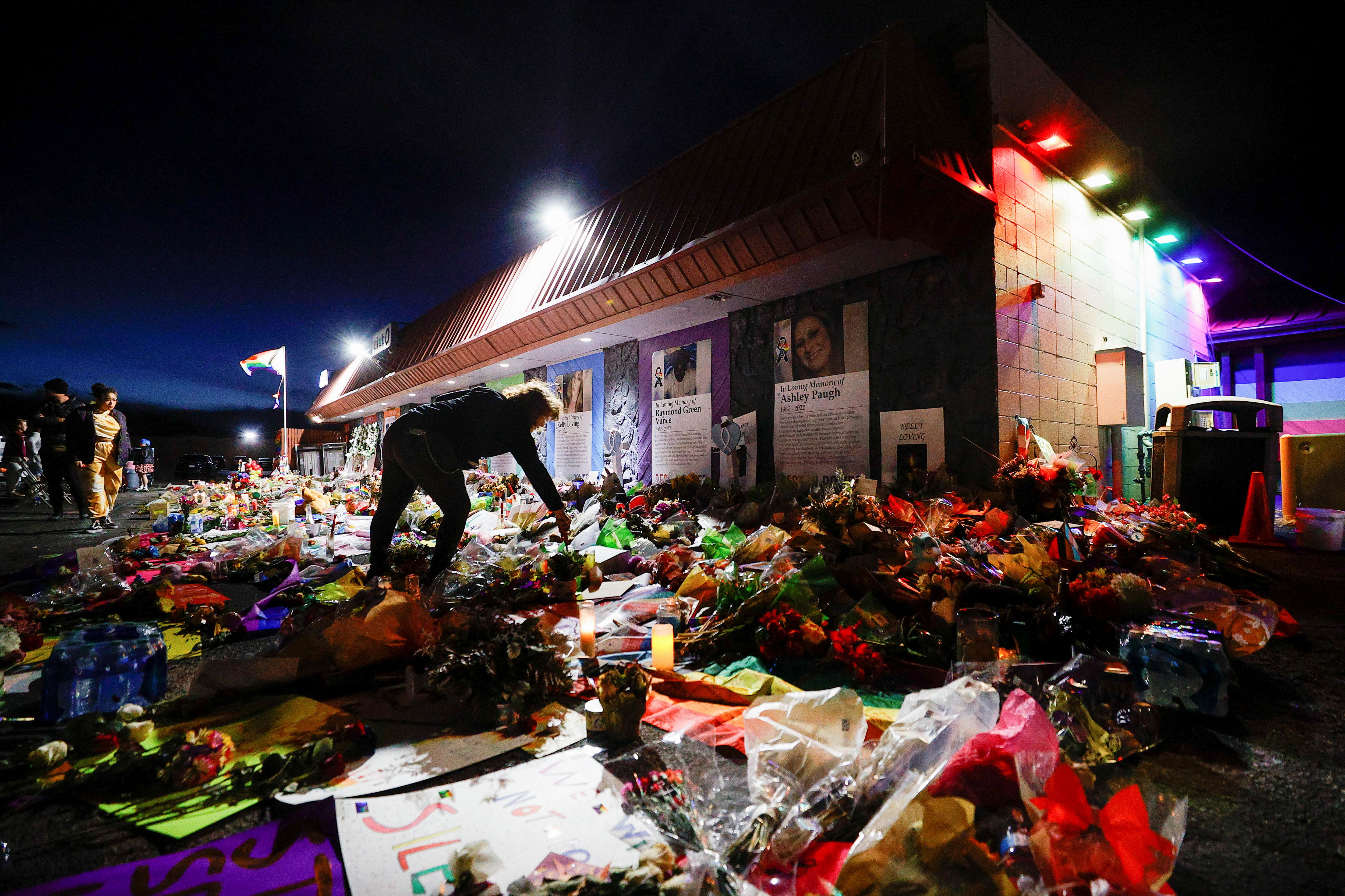A person lights a candle outside a nightclub, they are surrounded by floral tributes.