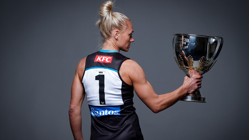 Erin Phillips, with her back to the camera, holds the Premiership Cup to her right and stares into it.
