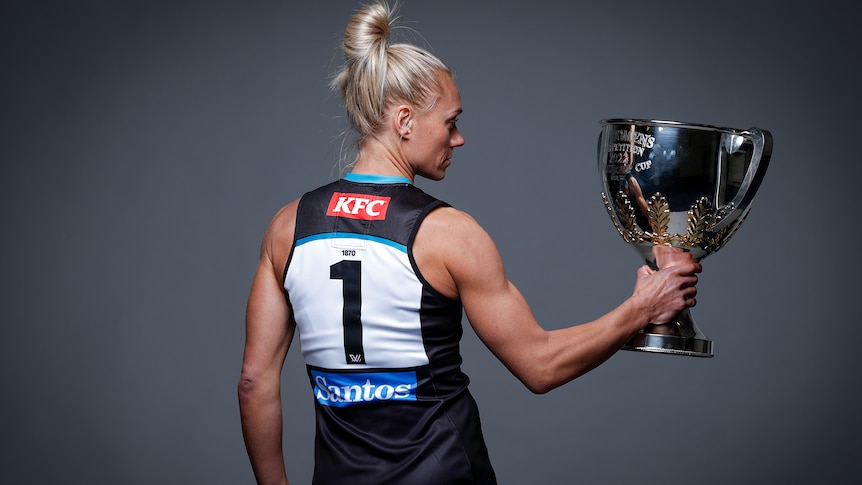 Erin Phillips, with her back to the camera, holds the Premiership Cup to her right and stares into it