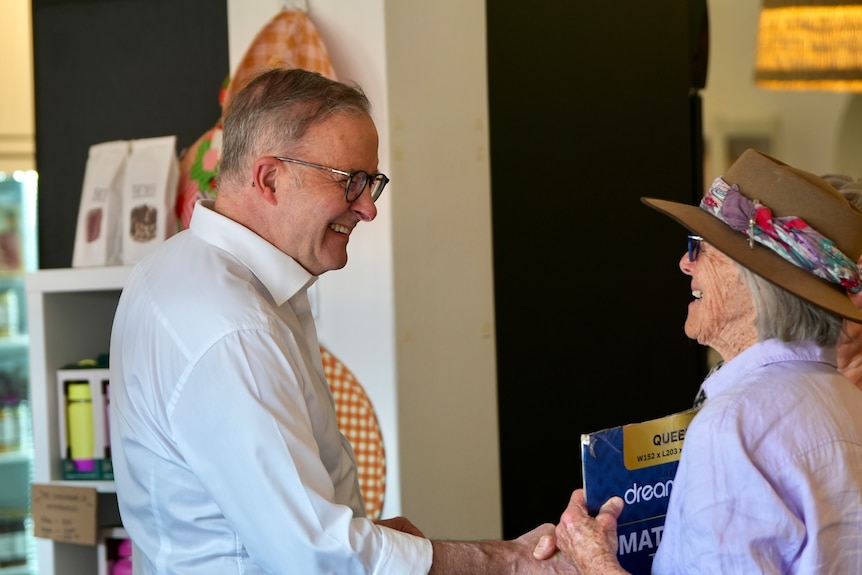 A smiling man in a white shirt and glasses shakes the hand of an older woman in a hat and glasses