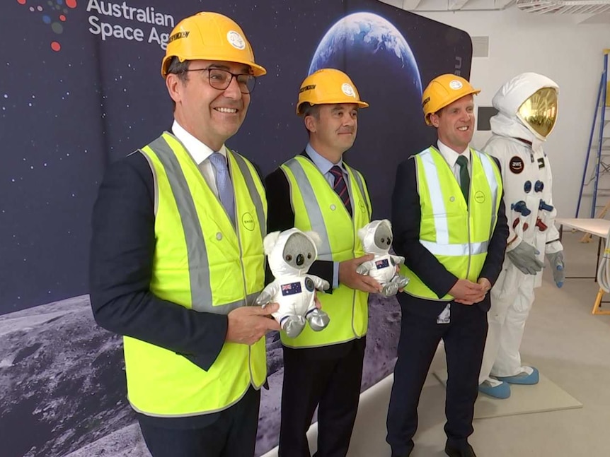 Three white men in hard hats stand holding stuffed toy koalas in space suits smiling for cameras at an event opening