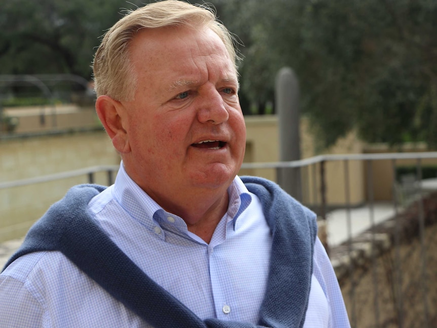 A man looks just beyond the camera. He is wearing a checked blue and white shirt with a dark jumper draped over his shoulders