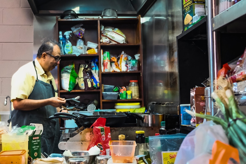 Un homme portant un tablier vu de profil à l'intérieur d'une cuisine, entouré d'ingrédients.