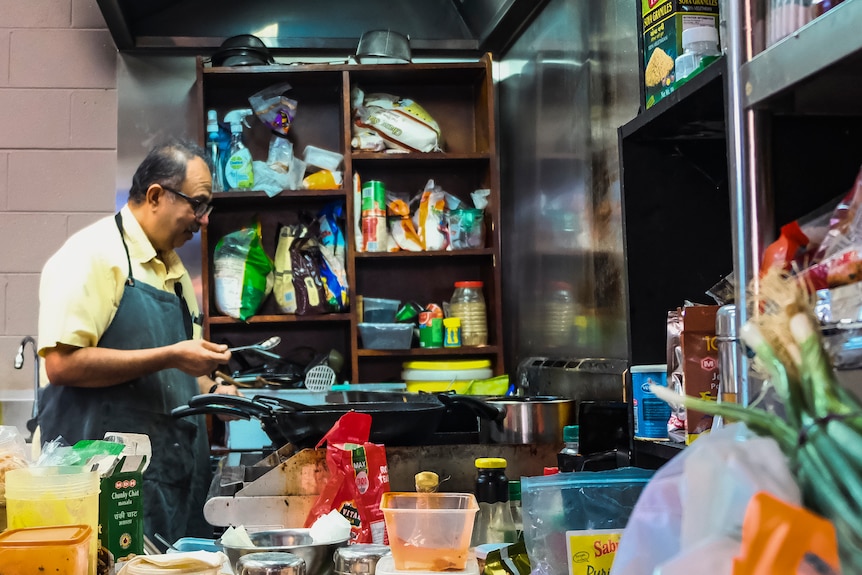 A man wearing an apron seen in side profile inside a kitchen, surrounded by ingredients.