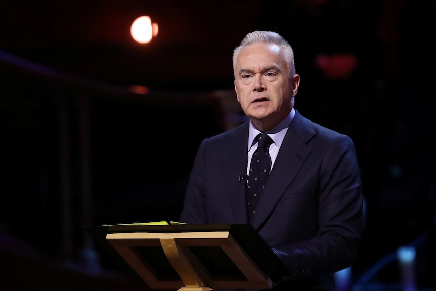 A man in a suit speaking at a lecturn, looking on.