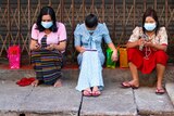 Three women in face masks sitting on a road side looking at their phones