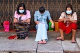 Three women in face masks sitting on a road side looking at their phones