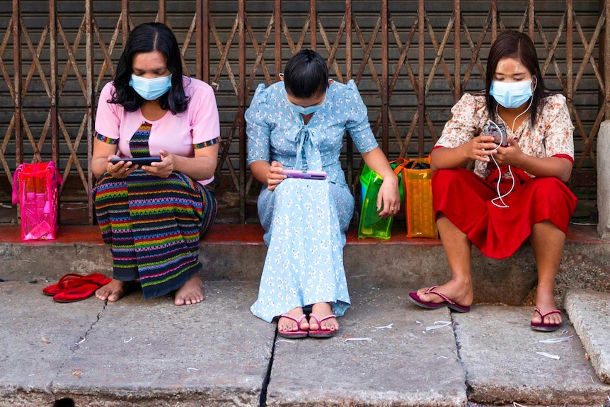 Three women in face masks sitting on a road side looking at their phones