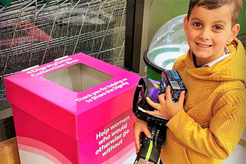 A young boy wearing a mustard jumper holding sanitary products above a donation bin smiling