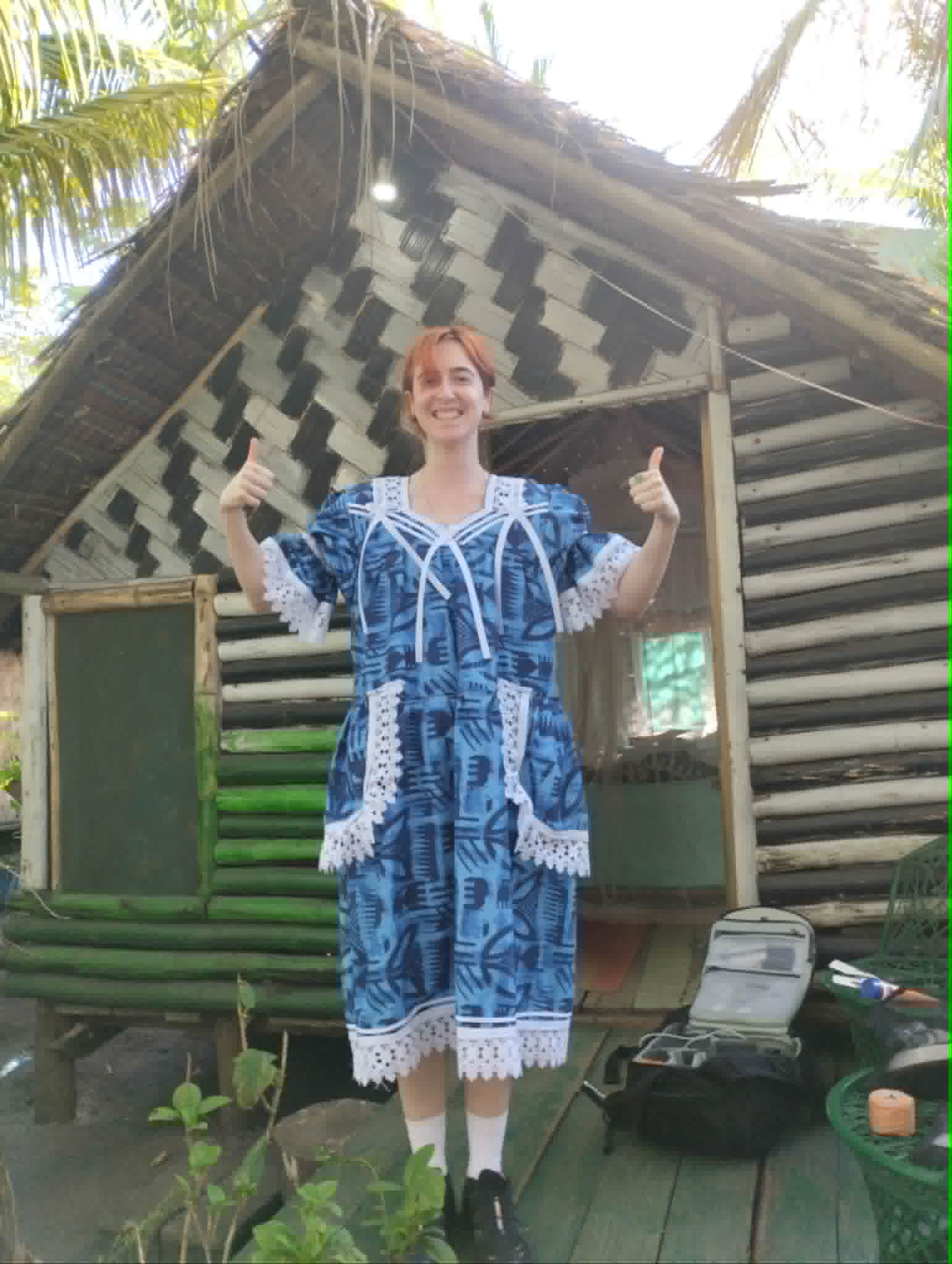 Kate wears a blue traditional dress from Vanuatu, holding her thumbs up and smiling.