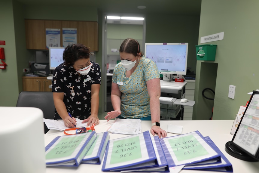 Two midwives in patterned scrubs stand at a table in a staff area, looking down at paperwork and bed folders.