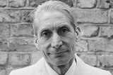 Headshot of Rolling Stones drummer Charlie Watts smiling at the camera. He stands before a brick wall. His hair is slicked back.