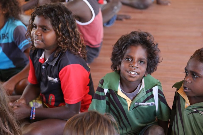 A group of young students smile and sit on a school hall floor.