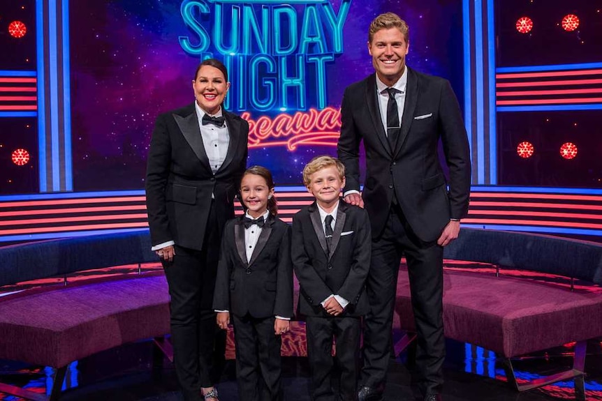 Chris Brown,  Julia Morris and two children all wear tuxedos on a set with the neon sign 'Saturday Night Takeaway' behind them.
