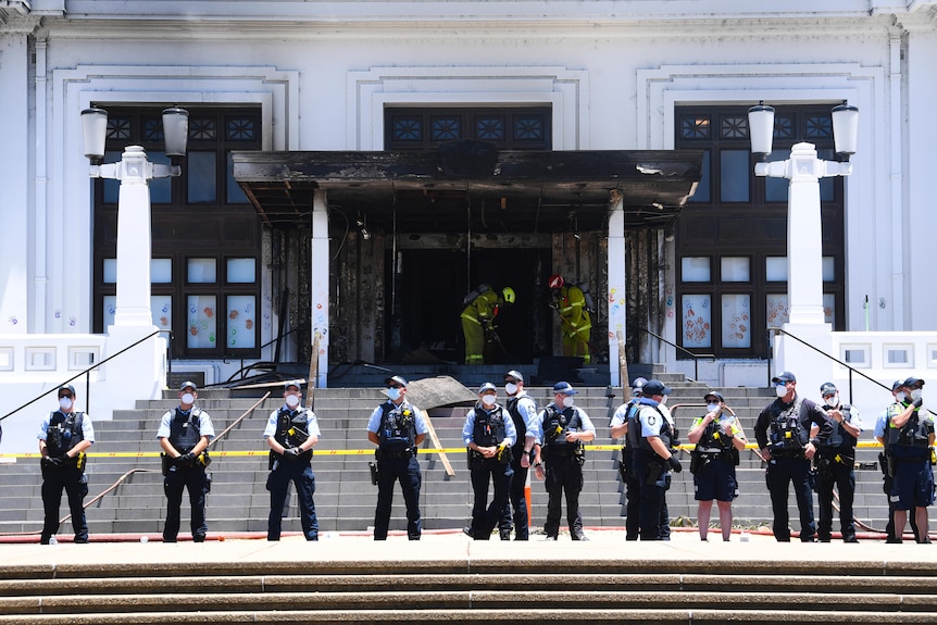 Police officers stand in a line in front of a burnt door.