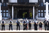 Police officers stand in a line in front of a burnt door.