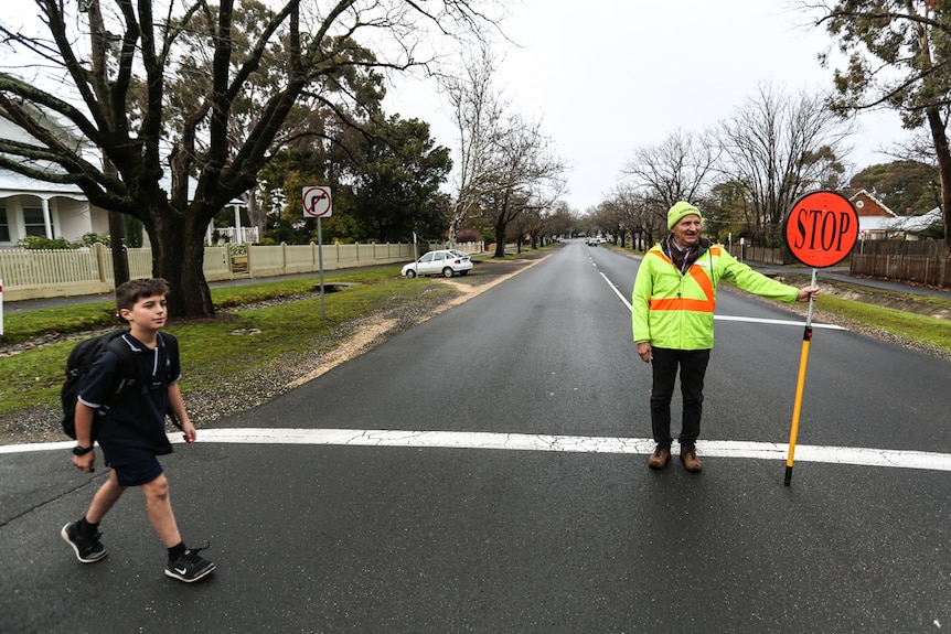 Schoolboy, dressed in blue uniform and with backpack, crosses the road. Crossing supervisor holds stop sign aloft.