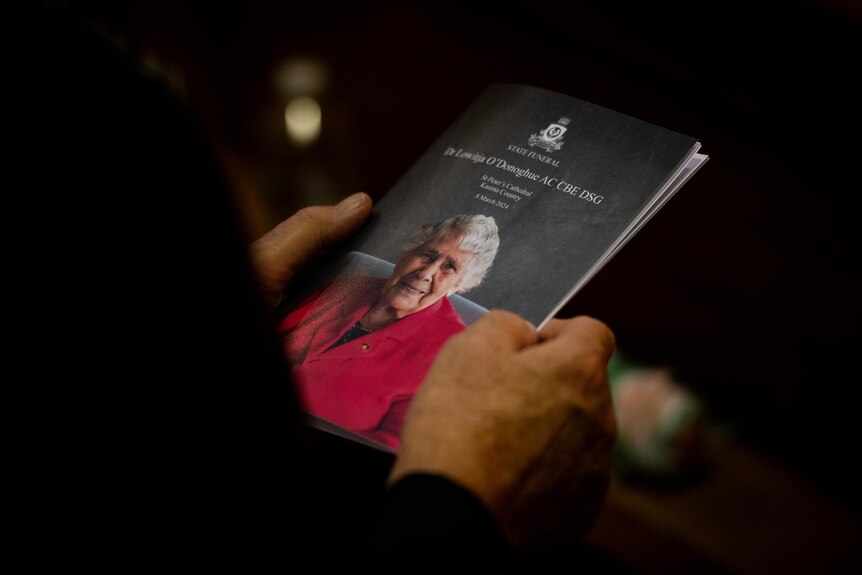 A close up of a person's hands holding a funeral program with an image of Dr Lowitja O'Donoghue on it