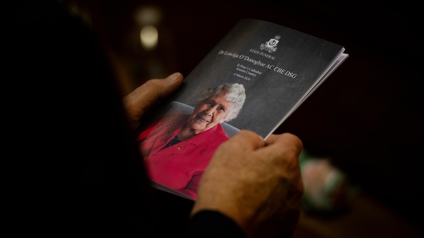 A close up of a person's hands holding a funeral program with an image of Dr Lowitja O'Donoghue on it