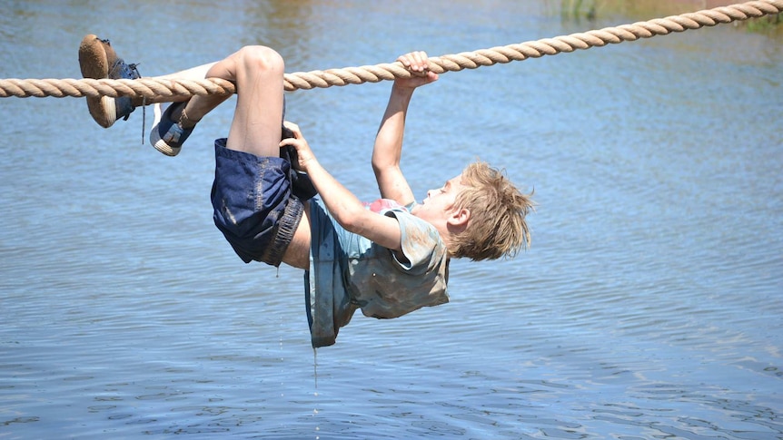 A boy grips a rope with one hand and both legs as he crosses a water obstacle.