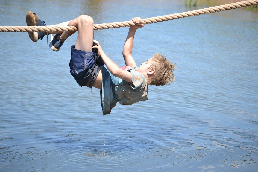 A boy grips a rope with one hand and both legs as he crosses a water obstacle.