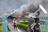 A broken plane is burning on the tarmac and some men are standing nearby.