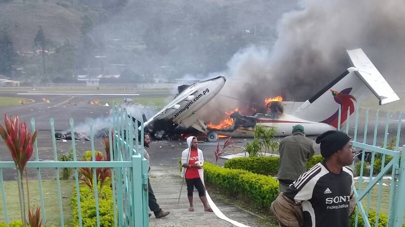 A broken plane is burning on the tarmac and some men are standing nearby.