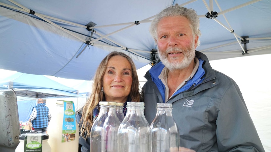 two farmers stand behind a row of glass bottles inside a tent