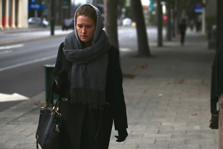 A woman wears a thick black jacket, gloves, and a scarf over her head in a cold morning on a city street.