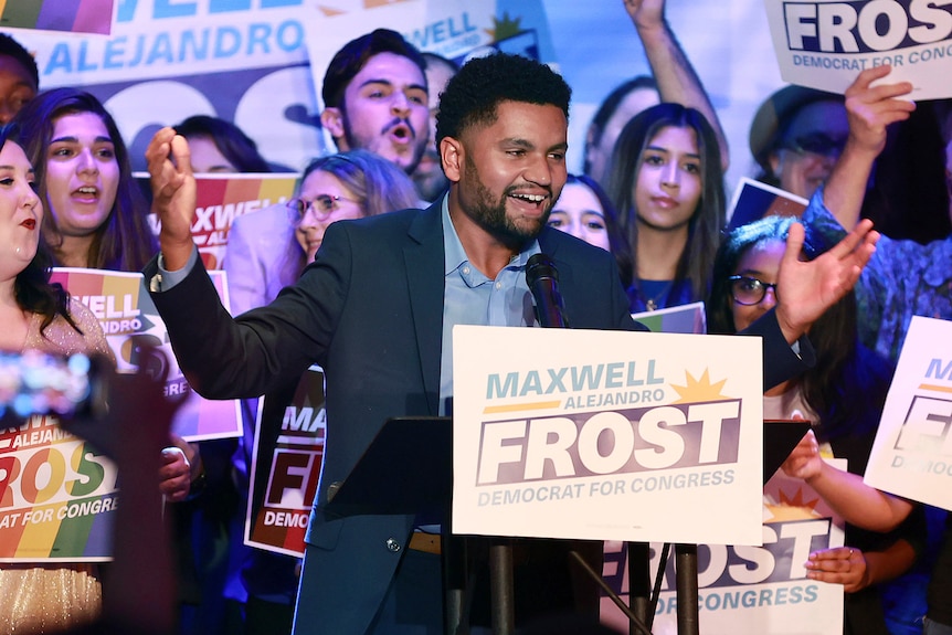 Maxwell standing at a podium with his supporters behind him. 