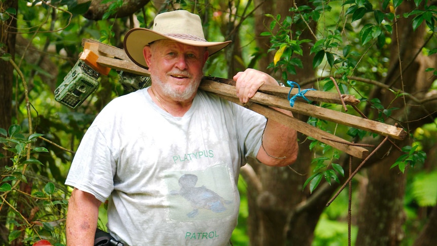 A man in his seventies holds wooden stakes with wildlife camera traps attached to them over his shoulder.