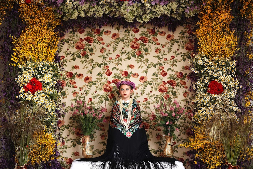 A young girl sits still in front of an altar decorated with colourful flowers.