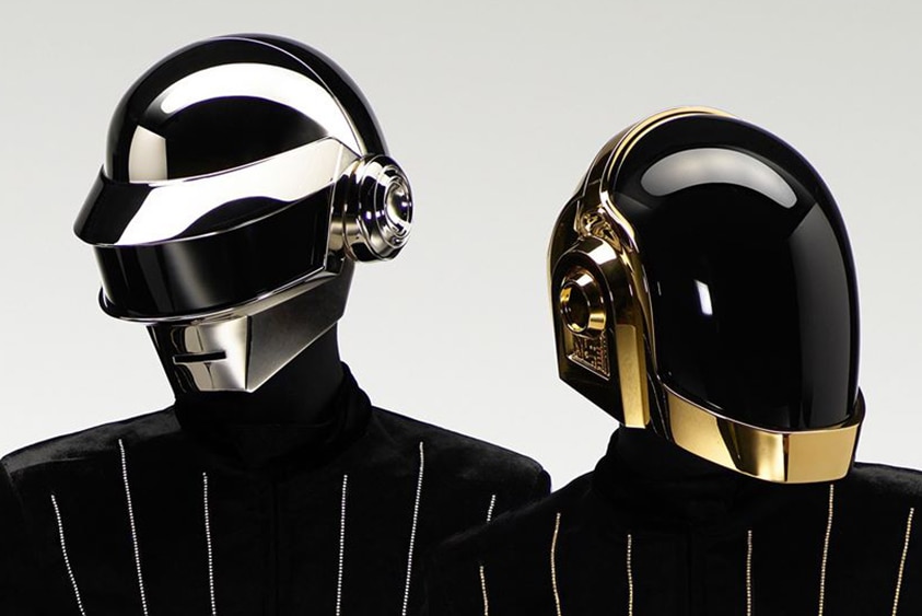Two members of Daft Punk wearing shiny silver and gold helmets and black suits