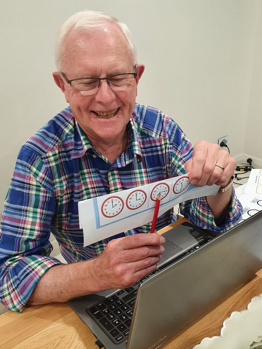 older man smiling, holding up paper with clocks telling the time in front of laptop screen