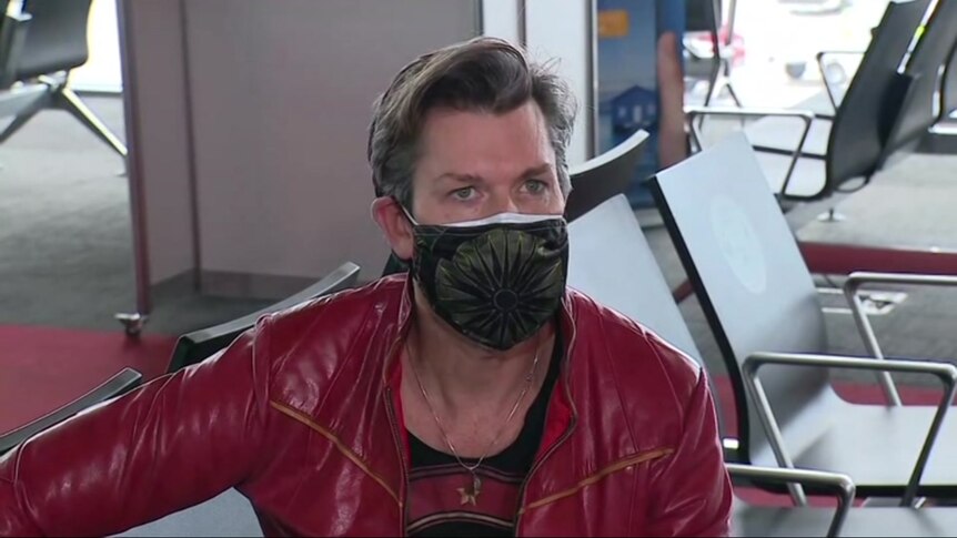 A man in a red jacket wears a mask while pictured seated in an airport lounge